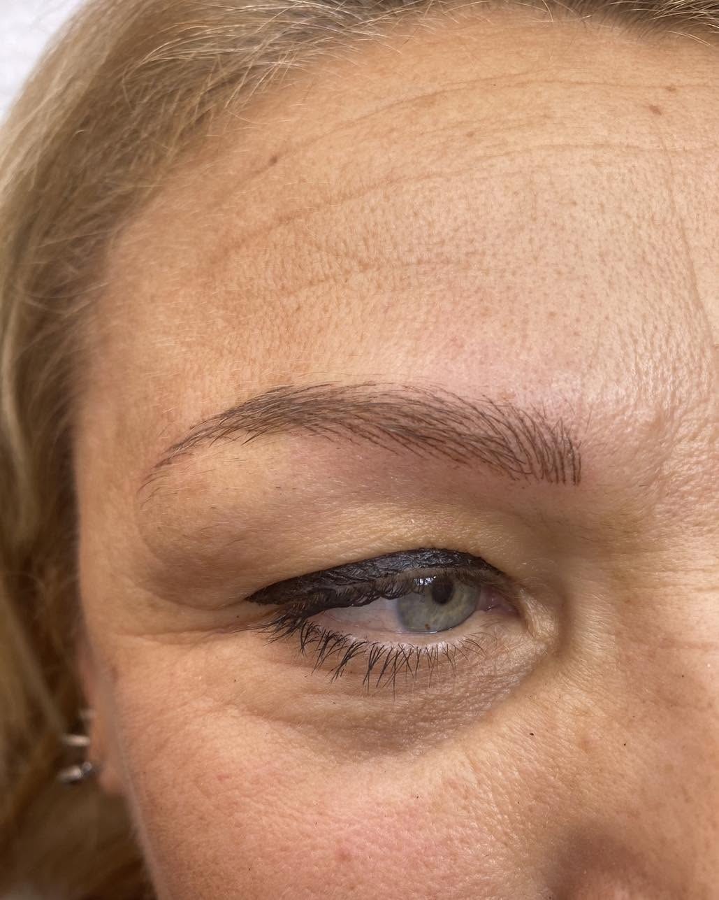 Sopracciglio castano disegnato a tratti sottili sopra un occhio verde con eyeliner nero, tra ciocche di capelli biondi